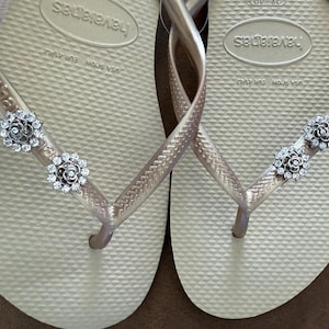 以下が含まれることがあります： ゴールドカラーの Havaianas ビーチサンダルのペア。シルバーのフローラル装飾が施されています。ビーチサンダルは、テクスチャード加工されたフットベッドと細いストラップを備えています。装飾は、小さく輝くクリスタルで作られています。