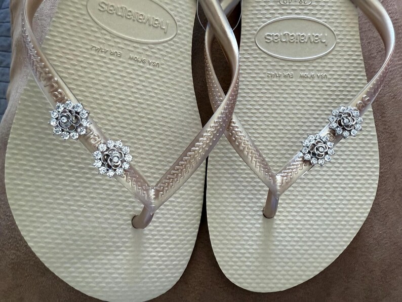 以下が含まれることがあります： ゴールドカラーの Havaianas ビーチサンダルのペア。シルバーのフローラル装飾が施されています。ビーチサンダルは、テクスチャード加工されたフットベッドと細いストラップを備えています。装飾は、小さく輝くクリスタルで作られています。