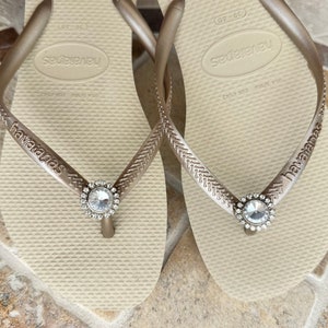 May include: A pair of beige Havaianas flip-flops with silver straps. Each strap has a clear, circular jewel. The brand name "havaianas" is printed on the strap.
