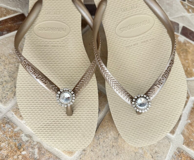 May include: A pair of beige Havaianas flip-flops with silver straps. Each strap has a clear, circular jewel. The brand name "havaianas" is printed on the strap.