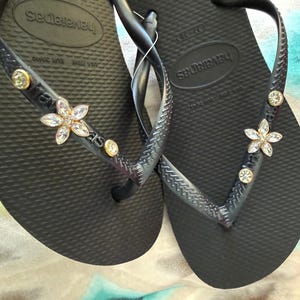 May include: Black Havaianas flip-flops with a textured footbed. The straps are embellished with a flower and round crystal accents. The flip-flops are a size 37-38.
