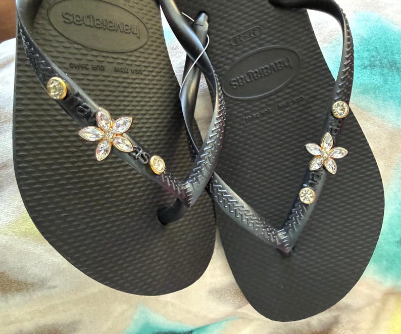 May include: Black Havaianas flip-flops with a textured footbed. The straps are embellished with a flower and round crystal accents. The flip-flops are a size 37-38.
