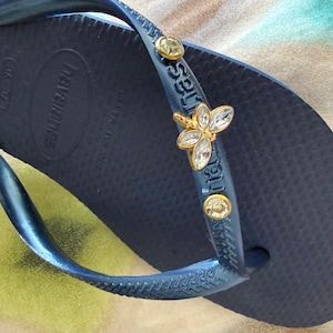 May include: Navy blue Havaianas flip-flop with a decorative gold and crystal butterfly charm on the strap. The flip-flop has a textured footbed and the brand name is visible on the side.