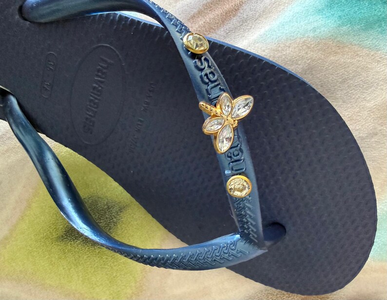 May include: Navy blue Havaianas flip-flop with a decorative gold and crystal butterfly charm on the strap. The flip-flop has a textured footbed and the brand name is visible on the side.