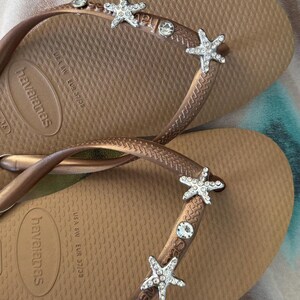 K&ouml;nnte beinhalten: Braune Havaianas-Flip-Flops mit dekorativen Seesternen und Strasssteinen. Die Flip-Flops haben ein strukturiertes Fu&szlig;bett und einen metallisch braunen Riemen. Gr&ouml;&szlig;e EUR 37/38.