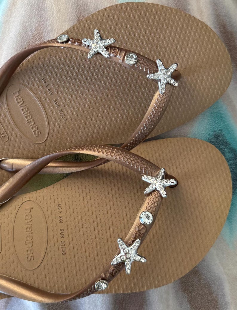 K&ouml;nnte beinhalten: Braune Havaianas-Flip-Flops mit dekorativen Seesternen und Strasssteinen. Die Flip-Flops haben ein strukturiertes Fu&szlig;bett und einen metallisch braunen Riemen. Gr&ouml;&szlig;e EUR 37/38.