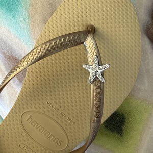 K&ouml;nnte beinhalten: Goldfarbene Havaianas-Zehensandale mit strukturierter Sohle und Riemen. Der Riemen hat einen silbernen Seestern-Charm. Die Flip-Flop-Gr&ouml;&szlig;e ist USA 7/8, EUR 39/40, Gr&ouml;&szlig;e 37-38. Die Sohle ist ca. 24 cm lang.