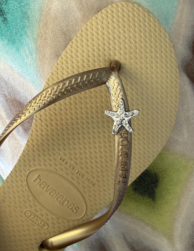 K&ouml;nnte beinhalten: Goldfarbene Havaianas-Zehensandale mit strukturierter Sohle und Riemen. Der Riemen hat einen silbernen Seestern-Charm. Die Flip-Flop-Gr&ouml;&szlig;e ist USA 7/8, EUR 39/40, Gr&ouml;&szlig;e 37-38. Die Sohle ist ca. 24 cm lang.