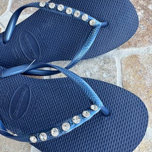 May include: A pair of navy blue flip-flops with a row of rhinestones on the strap. The flip-flops are made by Havaianas.