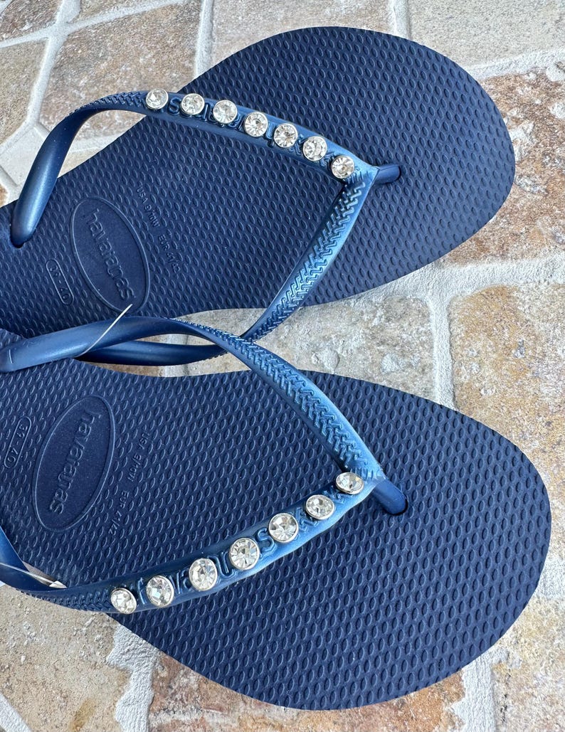 May include: A pair of navy blue flip-flops with a row of rhinestones on the strap. The flip-flops are made by Havaianas.