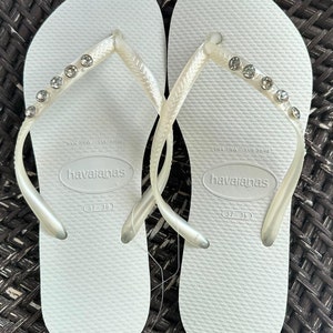 May include: A pair of white flip-flops with rhinestones on the straps. The flip-flops are labeled "havaianas" and have the size "37-38" printed on the bottom.