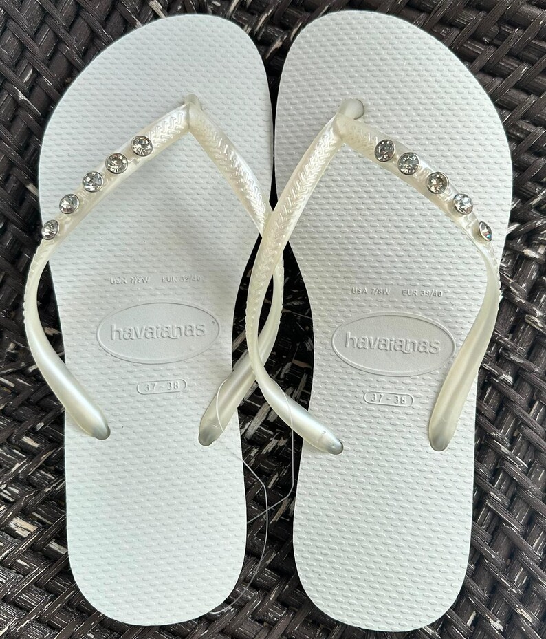 May include: A pair of white flip-flops with rhinestones on the straps. The flip-flops are labeled "havaianas" and have the size "37-38" printed on the bottom.