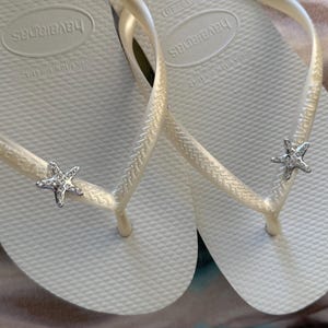 K&ouml;nnte beinhalten: Ein Paar wei&szlig;e Havaianas-Flip-Flops mit strukturierter Sohle und cremefarbenen Riemen. Jeder Riemen hat einen silbernen Seestern-Charm mit klaren Kristallakzenten. Der Markenname ist auf dem Fu&szlig;bett sichtbar.