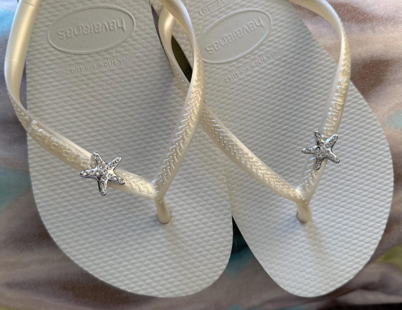 K&ouml;nnte beinhalten: Ein Paar wei&szlig;e Havaianas-Flip-Flops mit strukturierter Sohle und cremefarbenen Riemen. Jeder Riemen hat einen silbernen Seestern-Charm mit klaren Kristallakzenten. Der Markenname ist auf dem Fu&szlig;bett sichtbar.