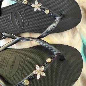 May include: Black Havaianas flip-flops with flower and gem embellishments. The sandals have a textured footbed and the brand name is visible on the strap and sole. The straps are decorated with a flower and several round gems.