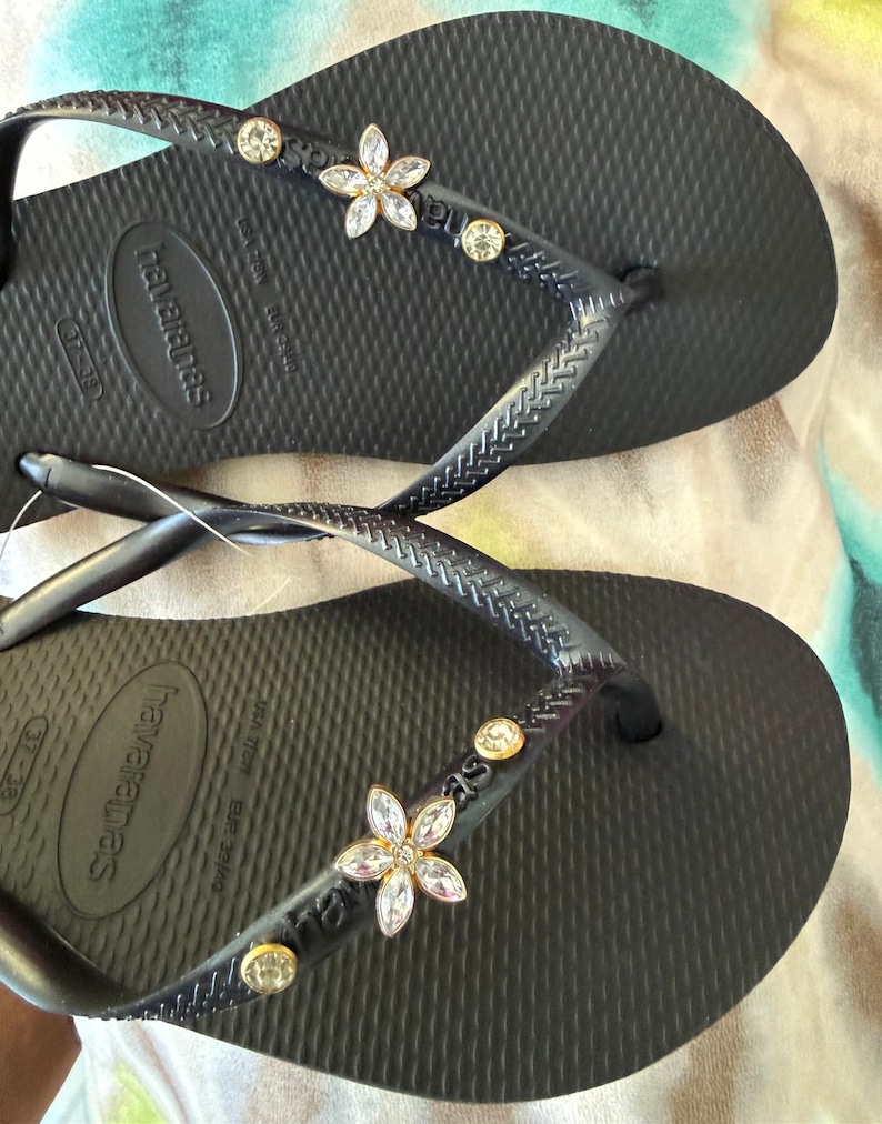 May include: Black Havaianas flip-flops with flower and gem embellishments. The sandals have a textured footbed and the brand name is visible on the strap and sole. The straps are decorated with a flower and several round gems.