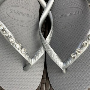 May include: A pair of gray flip-flops with rhinestones on the straps. The flip-flops have the word "havaianas" printed on the footbed.