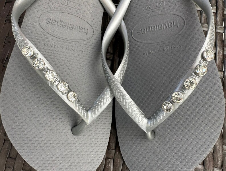 May include: A pair of gray flip-flops with rhinestones on the straps. The flip-flops have the word "havaianas" printed on the footbed.