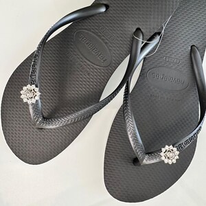 Pode incluir: Um par de chinelos pretos com um enfeite de strass prateado em cada tira. Os chinelos s&atilde;o etiquetados com "Havaianas" na sola.