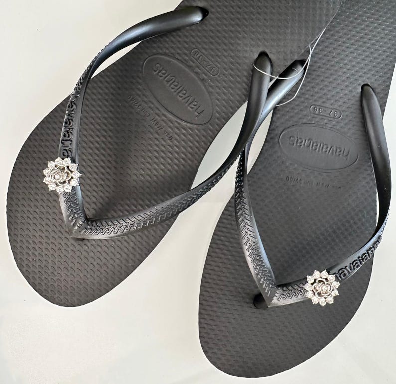 Pode incluir: Um par de chinelos pretos com um enfeite de strass prateado em cada tira. Os chinelos s&atilde;o etiquetados com "Havaianas" na sola.