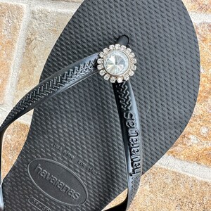 May include: Black Havaianas flip-flop with a decorative rhinestone embellishment. The flip-flop has a textured surface and the brand name is visible on the strap. The embellishment is round with a large central stone and smaller surrounding stones.