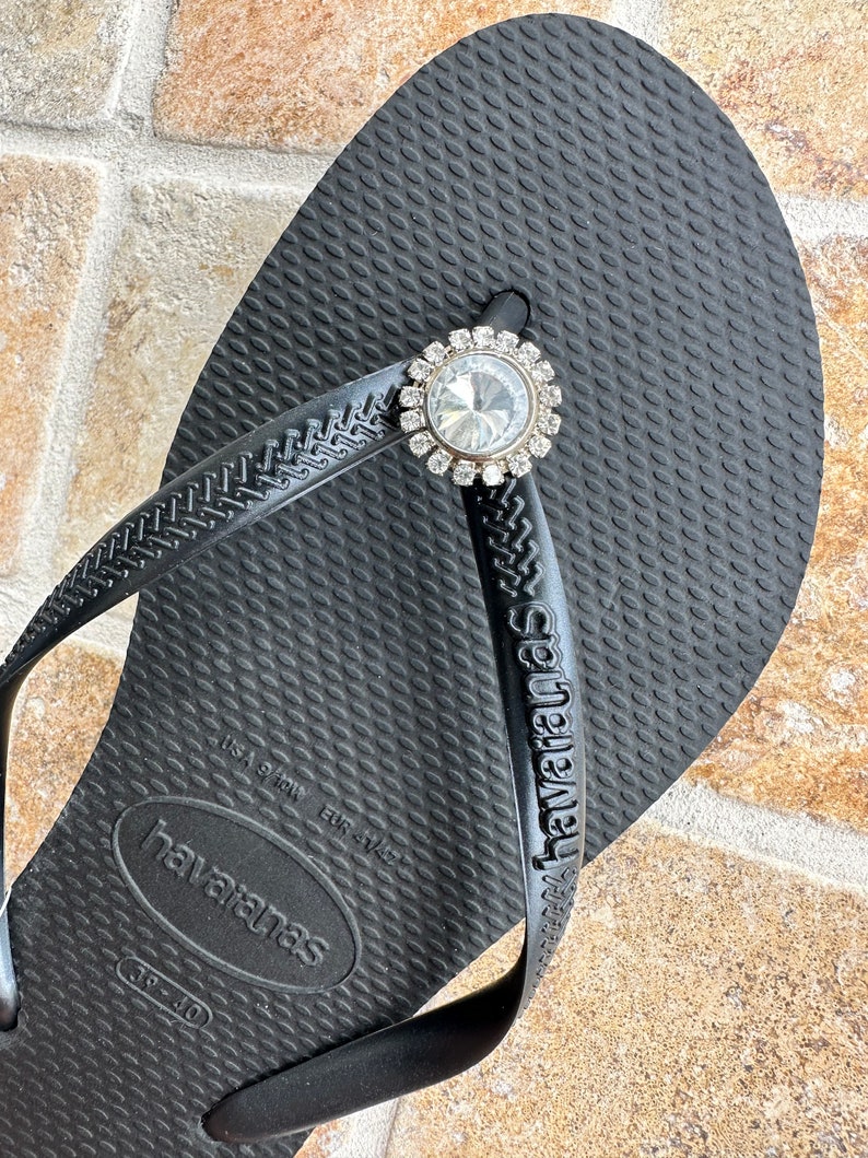 May include: Black Havaianas flip-flop with a decorative rhinestone embellishment. The flip-flop has a textured surface and the brand name is visible on the strap. The embellishment is round with a large central stone and smaller surrounding stones.