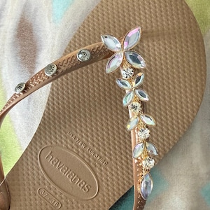 May include: A tan Havaianas flip-flop with a decorative floral design. The strap features clear, round rhinestones and a gold-colored metal accent. The floral design is made of iridescent, petal-shaped rhinestones.
