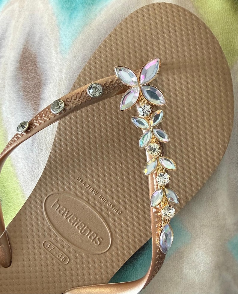 May include: A tan Havaianas flip-flop with a decorative floral design. The strap features clear, round rhinestones and a gold-colored metal accent. The floral design is made of iridescent, petal-shaped rhinestones.