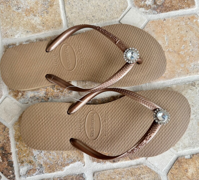 May include: A pair of brown Havaianas flip-flops with embellished straps. Each strap has a round, clear jewel. The flip-flops are tan with a textured footbed and the Havaianas logo.