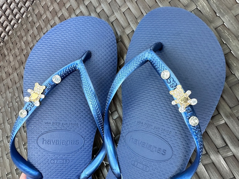 Puede incluir: Un par de chanclas azules con un adorno dorado y de pedrer&iacute;a en cada tira. Las chanclas est&aacute;n etiquetadas como "havaianas" y tienen la talla "37-38" impresa en la parte inferior.