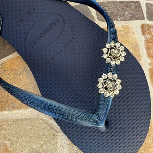 以下が含まれることがあります： ネイビーブルーのハワイアナスビーチサンダル。ストラップには、花型のラインストーン装飾が2つ付いています。ビーチサンダルは、テクスチャード加工されたフットベッドと濃い青色のストラップが付いています。装飾は、クリアなラインストーンが付いたシルバーカラーです。