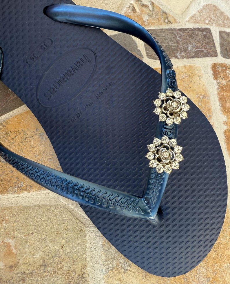 以下が含まれることがあります： ネイビーブルーのハワイアナスビーチサンダル。ストラップには、花型のラインストーン装飾が2つ付いています。ビーチサンダルは、テクスチャード加工されたフットベッドと濃い青色のストラップが付いています。装飾は、クリアなラインストーンが付いたシルバーカラーです。