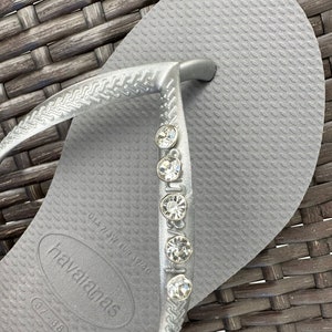 May include: Silver flip-flop sandals with a row of rhinestones on the strap. The sandals have the brand name "havaianas" and the size "37-38" printed on the sole.