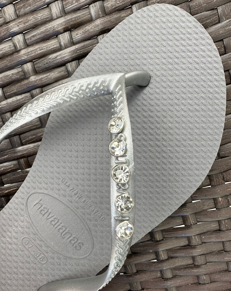 May include: Silver flip-flop sandals with a row of rhinestones on the strap. The sandals have the brand name "havaianas" and the size "37-38" printed on the sole.