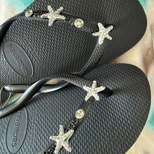 K&ouml;nnte beinhalten: Schwarze Havaianas-Flip-Flops mit strukturierter Sohle. Die Riemen sind mit silbernen Seesternen und klaren Strasssteinen verziert. Die Flip-Flops liegen auf einer gemusterten Oberfl&auml;che.
