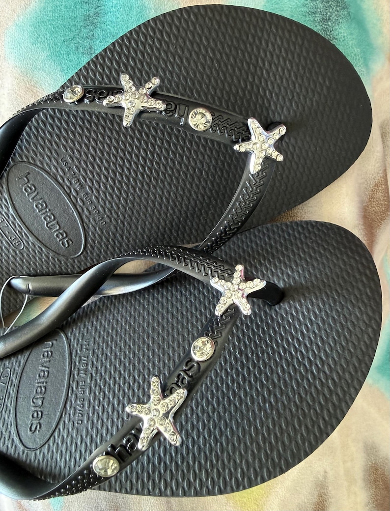 K&ouml;nnte beinhalten: Schwarze Havaianas-Flip-Flops mit strukturierter Sohle. Die Riemen sind mit silbernen Seesternen und klaren Strasssteinen verziert. Die Flip-Flops liegen auf einer gemusterten Oberfl&auml;che.
