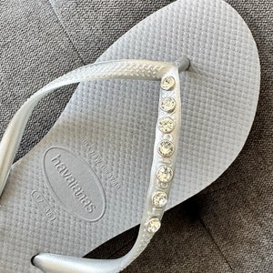 May include: Silver flip-flops with a row of rhinestones on the strap. The flip-flops have the brand name "havaianas" and the size "(37/38)" printed on the sole.