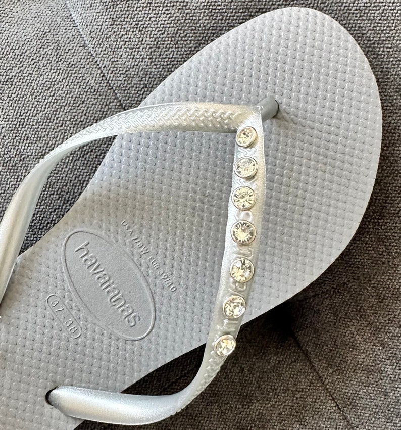 May include: Silver flip-flops with a row of rhinestones on the strap. The flip-flops have the brand name "havaianas" and the size "(37/38)" printed on the sole.