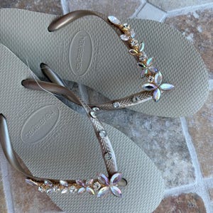 May include: A pair of beige Havaianas flip-flops with gold straps. The straps are adorned with a floral design of iridescent crystals and rhinestones. The flip-flops are displayed on a textured surface.