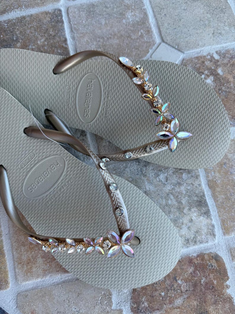 May include: A pair of beige Havaianas flip-flops with gold straps. The straps are adorned with a floral design of iridescent crystals and rhinestones. The flip-flops are displayed on a textured surface.