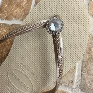 May include: Tan Havaianas flip-flop with a decorative silver and clear crystal embellishment. The flip-flop features a textured footbed and the brand name embossed on the strap. Size EUR 41/42.