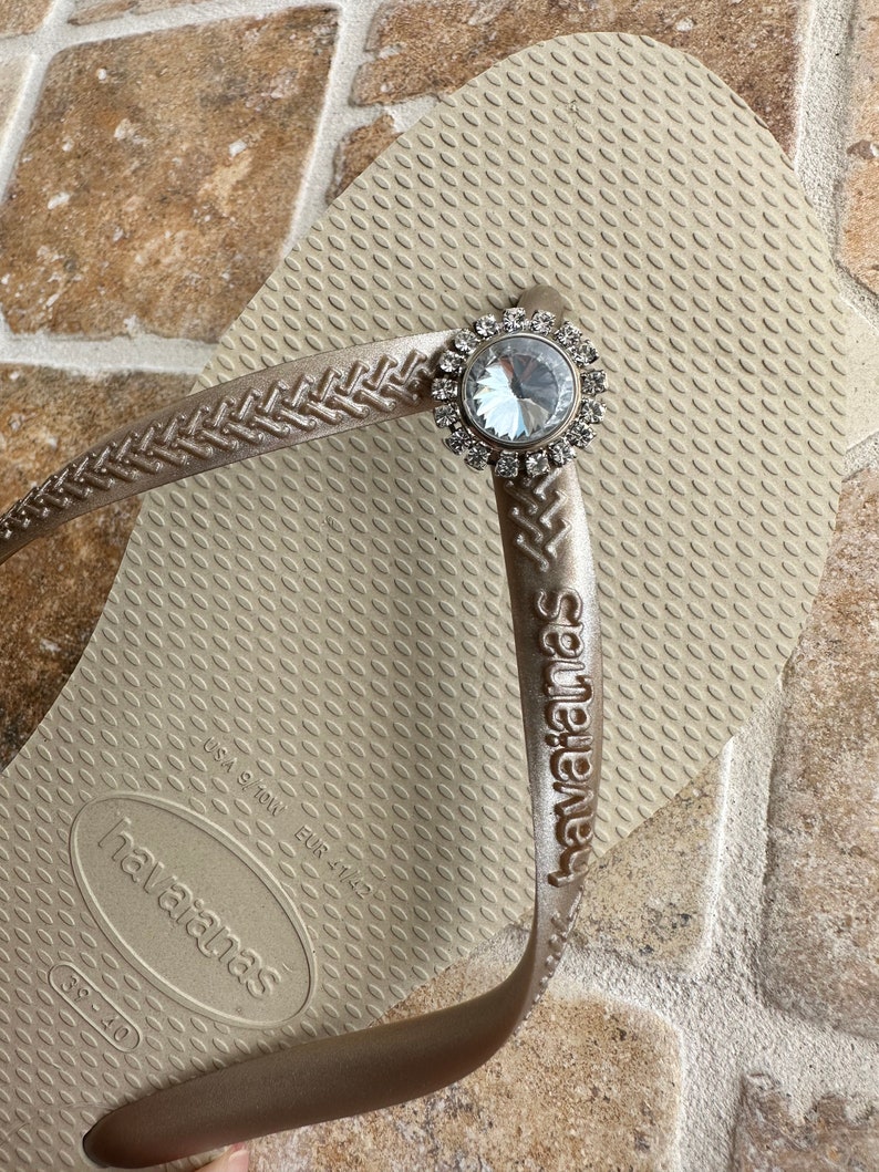 May include: Tan Havaianas flip-flop with a decorative silver and clear crystal embellishment. The flip-flop features a textured footbed and the brand name embossed on the strap. Size EUR 41/42.