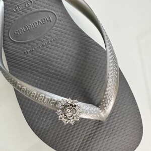 Pode incluir: Sand&aacute;lias Havaianas prateadas com um charme de flor prateada. As sand&aacute;lias t&ecirc;m o nome da marca "havaianas" impresso na tira.
