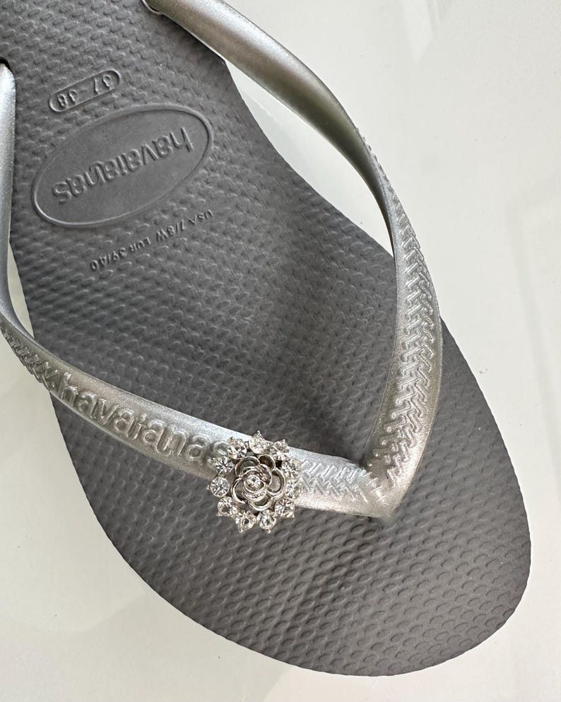 Pode incluir: Sand&aacute;lias Havaianas prateadas com um charme de flor prateada. As sand&aacute;lias t&ecirc;m o nome da marca "havaianas" impresso na tira.