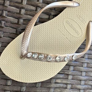 May include: A pair of beige flip-flops with a gold strap and rhinestones. The flip-flops have the brand name "havaianas" on the strap and the text "36-37" and "Brazil 38/39 MS 151" on the sole.