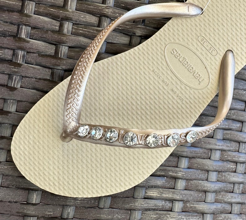 May include: A pair of beige flip-flops with a gold strap and rhinestones. The flip-flops have the brand name "havaianas" on the strap and the text "36-37" and "Brazil 38/39 MS 151" on the sole.