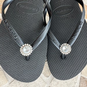 May include: A pair of black Havaianas flip-flops with a textured surface. Each flip-flop has a sparkling, round, clear rhinestone embellishment at the toe post. The brand name "Havaianas" is printed on the strap.