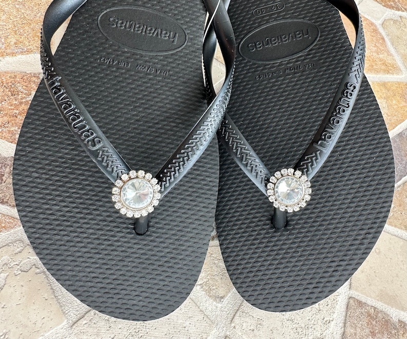 May include: A pair of black Havaianas flip-flops with a textured surface. Each flip-flop has a sparkling, round, clear rhinestone embellishment at the toe post. The brand name "Havaianas" is printed on the strap.
