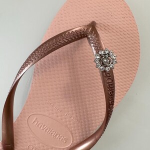 Pode incluir: Um par de chinelos rosa dourado com uma pequena flor de strass como enfeite na tira. Os chinelos s&atilde;o marcados "havaianas" e t&ecirc;m o tamanho "37-38" impresso na sola.