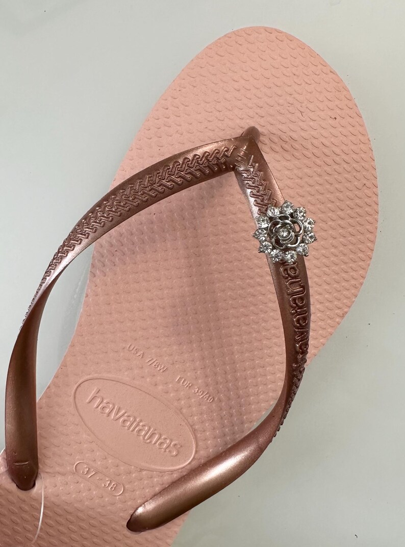 Pode incluir: Um par de chinelos rosa dourado com uma pequena flor de strass como enfeite na tira. Os chinelos s&atilde;o marcados "havaianas" e t&ecirc;m o tamanho "37-38" impresso na sola.